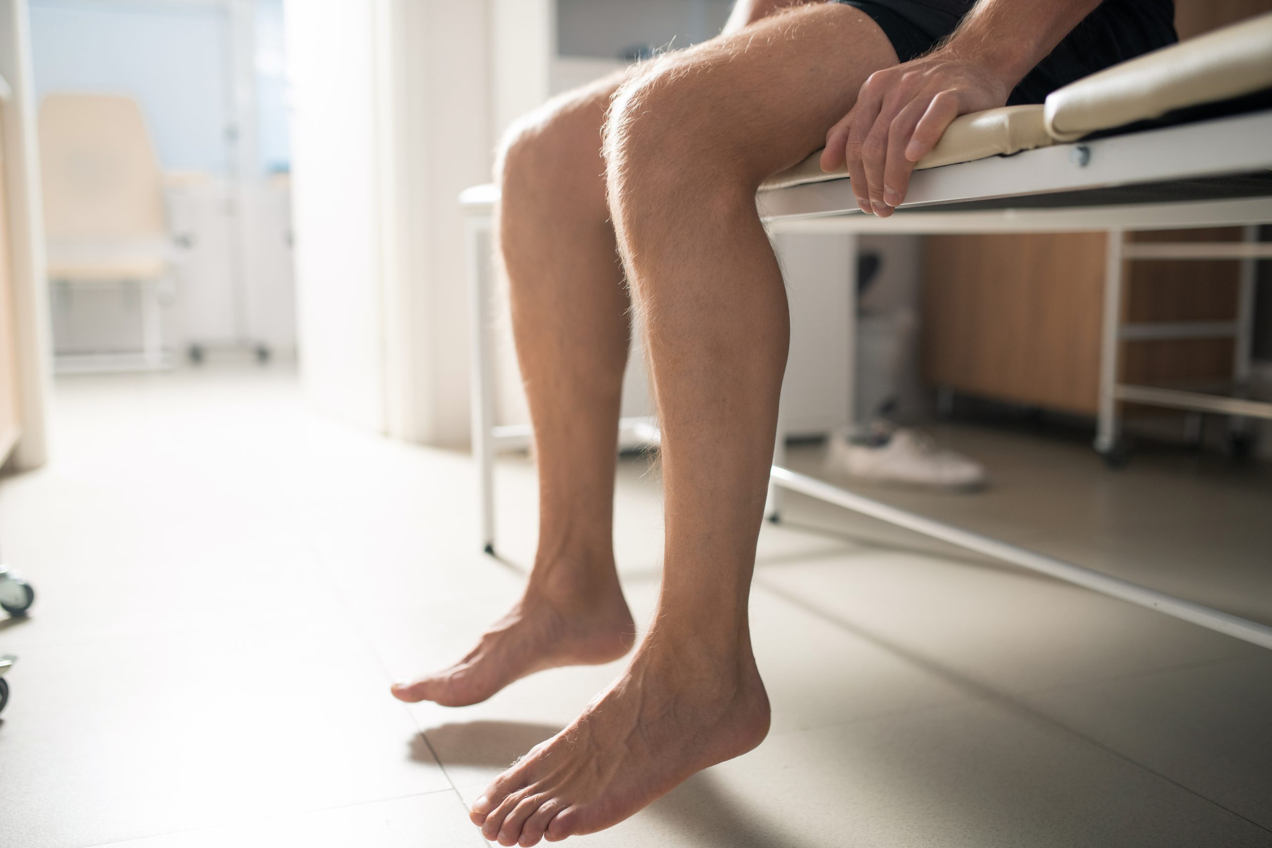 Bare legs of sick male patient sitting on couch in medical office| Featured Image for Identifying Flat Feet and Ankle Pronation | Blog