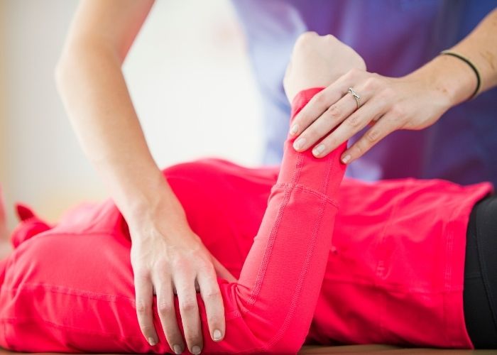 A close up of a woman's torso laying down while another person is bending their elbow and holding their bicep | Featured image for Throwers Elbow.