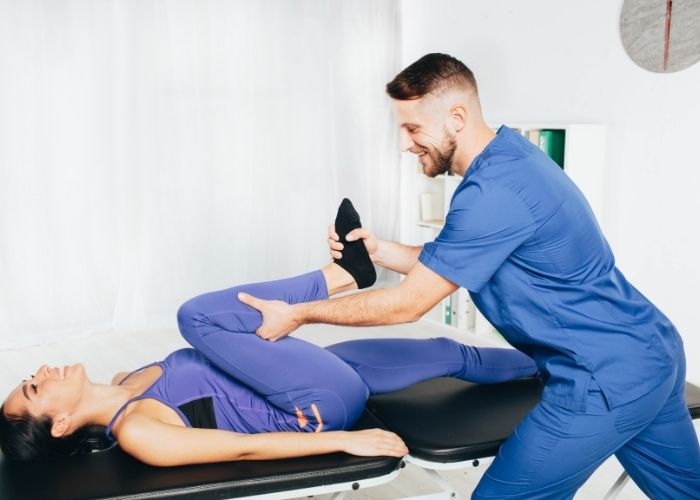 A woman laying on her back on a physio bed with one knee pressed into her chest and a man is stretching it out | Featured image for Hip Physiotherapy.