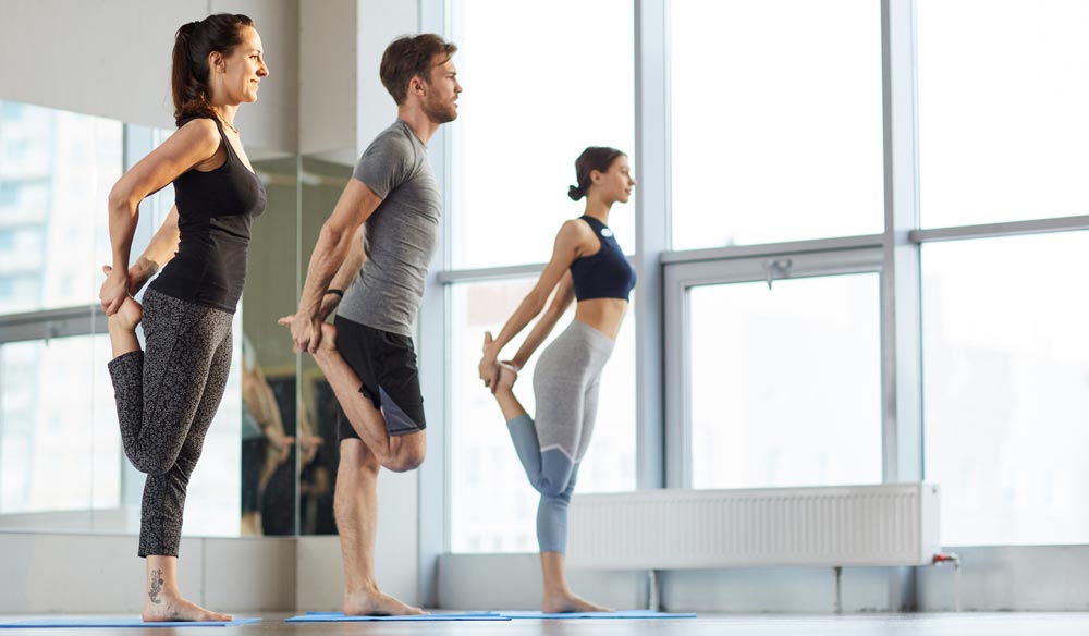 Three people standing on yoga mats holding one of their legs and stretching | Featured image on Osteitis Pubis.