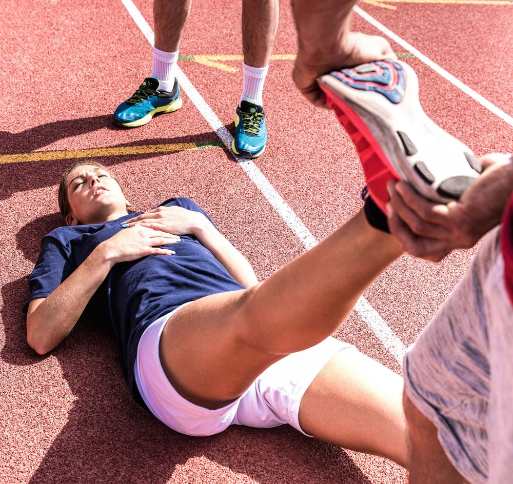 A person laying on their back on the floor on a track with their leg extended and another person stretching it out | Featured image on Hamstring Injury.