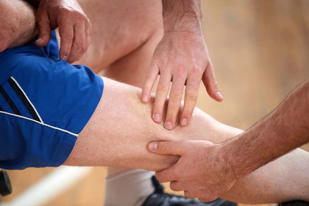 A close up of a persons knee with another person pressing their hand onto their knee | Featured image on Hamstring injury.