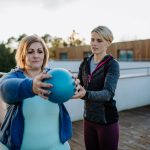 Woman exercising with a ball while being helped by a trainer | Featured image for the Blood Pressure Exercises page at Pivotal Motion Physiotherapy.