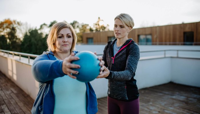 Woman exercising with a ball while being helped by a trainer | Featured image for the Blood Pressure Exercises page at Pivotal Motion Physiotherapy.